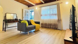 A living room with a wooden ceiling, a grey sofa with yellow cushions, a TV stand, and a window with floral curtains.