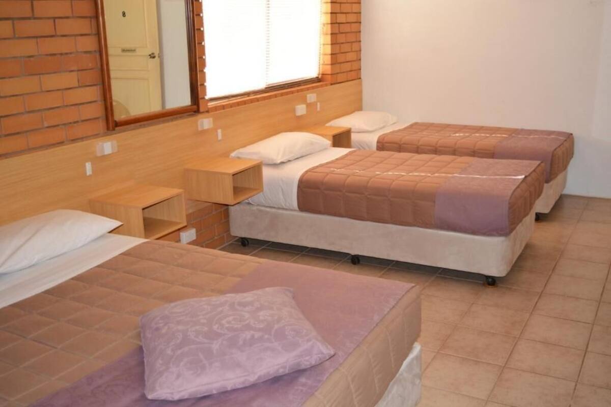 A hotel room with three beds, each with a wooden headboard, white bedding, and a small bedside table.