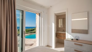A bedroom with a large window, a view of the sea, a wooden vanity, and a bed.