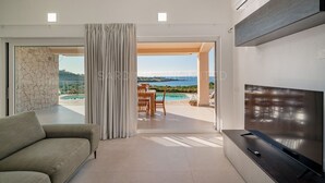 A modern living room with a sofa, a TV, and a view of a pool and landscape.