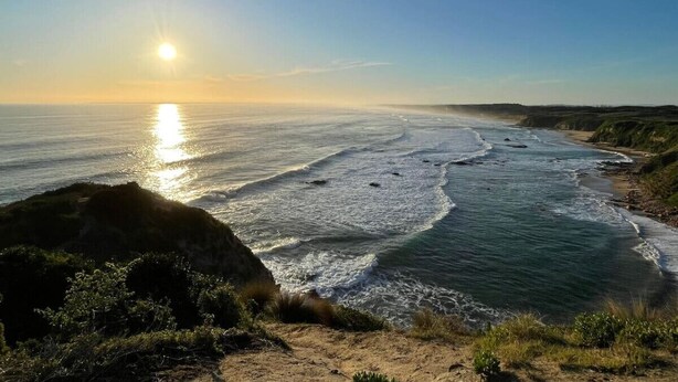 Paisagem costeira com céu claro, oceano iluminado pelo sol e uma praia arenosa.