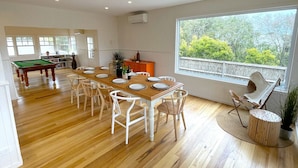 Uma sala de jantar com uma mesa de madeira posta para seis pessoas, rodeada por cadeiras brancas. Uma mesa de bilhar é visível ao fundo.