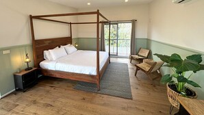 Uma cama com dossel em um quarto com piso de madeira, uma mesinha de cabeceira com luminária, uma grande planta em vaso e vista para o exterior por uma porta deslizante.