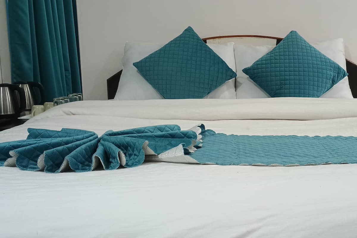 A neatly made bed with a teal quilted blanket and pillows, a bedside table with a kettle and cups, and a power outlet on the wall.