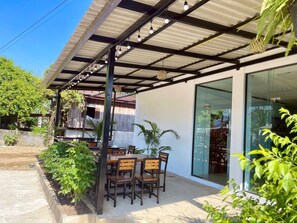 An outdoor patio with a table and chairs, surrounded by greenery and a covered area with hanging lights.
