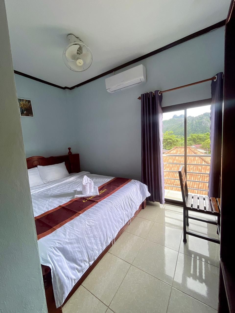 A hotel room with a bed, a chair, a window with curtains, an air conditioning unit, and a view of the outside.