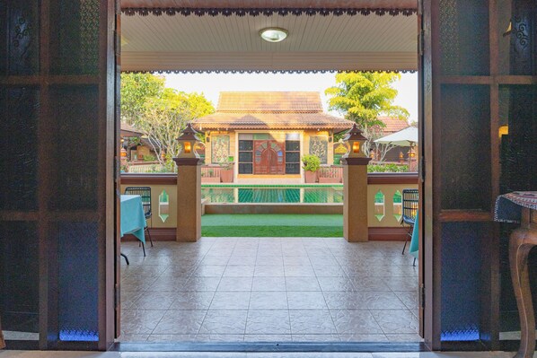 An open doorway leading to a covered patio area with tiled flooring, a green lawn, and a building with a tiled roof in the background. - Bannkrukaihomestay (San Sai)