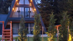 A modern A-frame cabin with a balcony, surrounded by trees and a wooden deck.