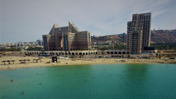 Zona costera con playa, un hotel grande de varios pisos y un paisaje montañoso al fondo.