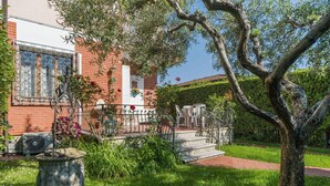 Une maison avec une façade en briques, un balcon blanc et un jardin agrémenté d’un arbre et d’un bac à fleurs en pierre.