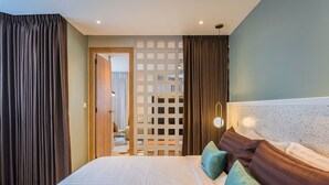 Une chambre d’hôtel avec un grand lit, deux oreillers, une tête de lit au motif texturé et une porte en bois donnant sur une autre pièce.