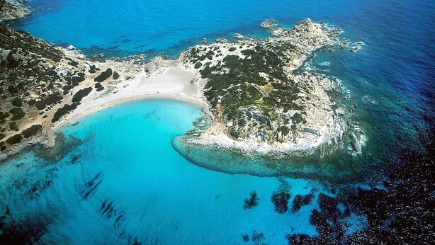 Vue aérienne d’une zone côtière avec une plage de sable, des eaux d’un bleu limpide et un terrain rocheux.