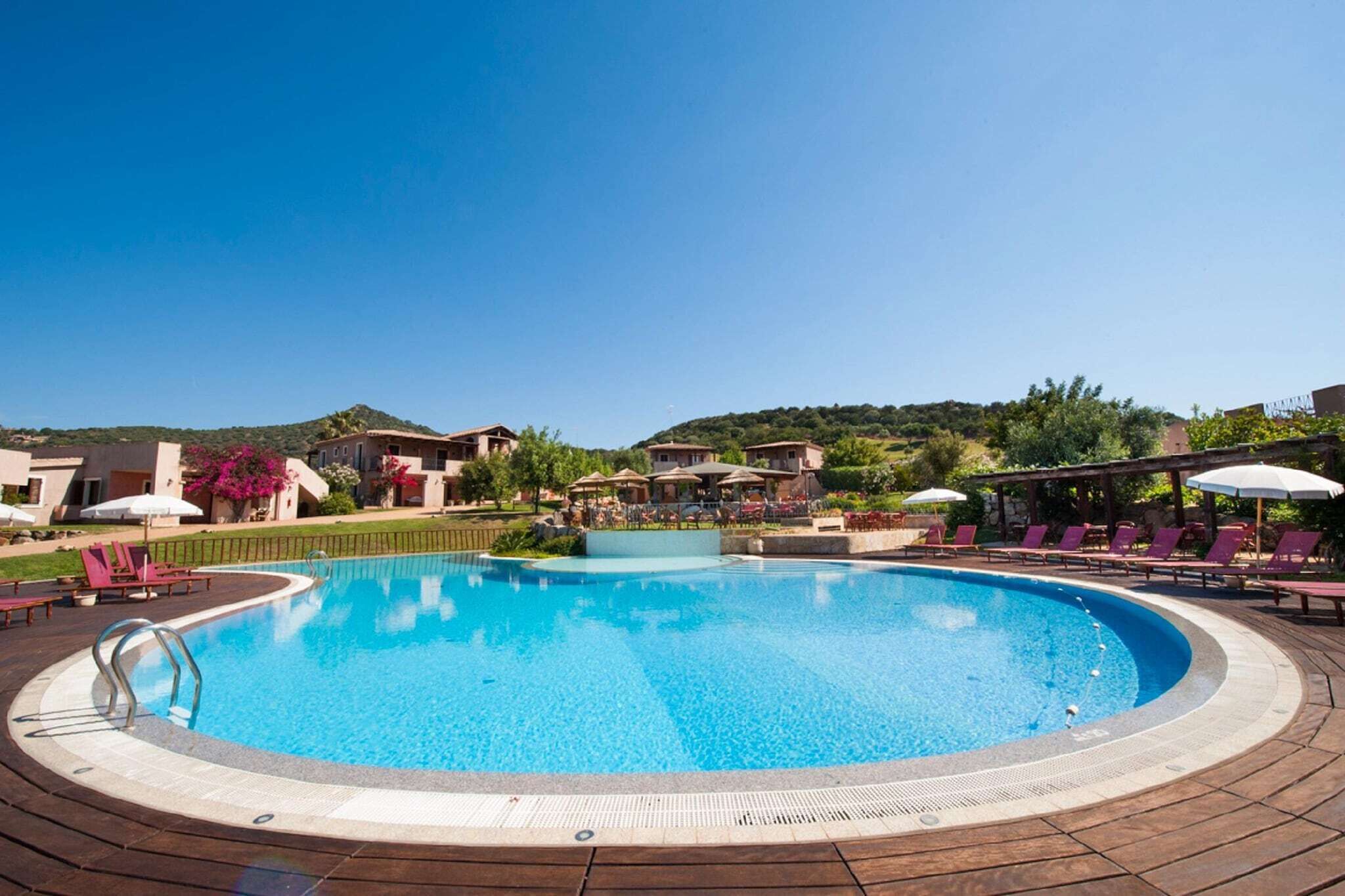 Ein großer, klarer Pool mit Liegestühlen und Sonnenschirmen, umgeben von begrünten Flächen und Gebäuden.