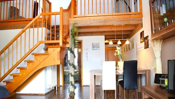 A spacious living area with a wooden staircase, a dining table with chairs, and a kitchen area visible through an open door. - Apartment Near Kurpark Lake (Bodenmais)