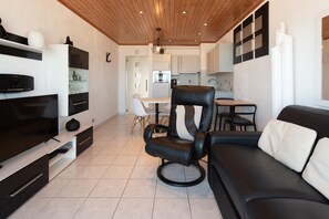 Un salon moderne avec un canapé en cuir noir, un fauteuil en cuir noir, un téléviseur à écran plat, une table à manger avec des chaises, et une cuisine en arrière-plan.