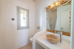 Une salle de bain avec un lavabo blanc, un récipient en bois servant de cuvette, un miroir avec un cadre décoratif et une fenêtre avec un store blanc.