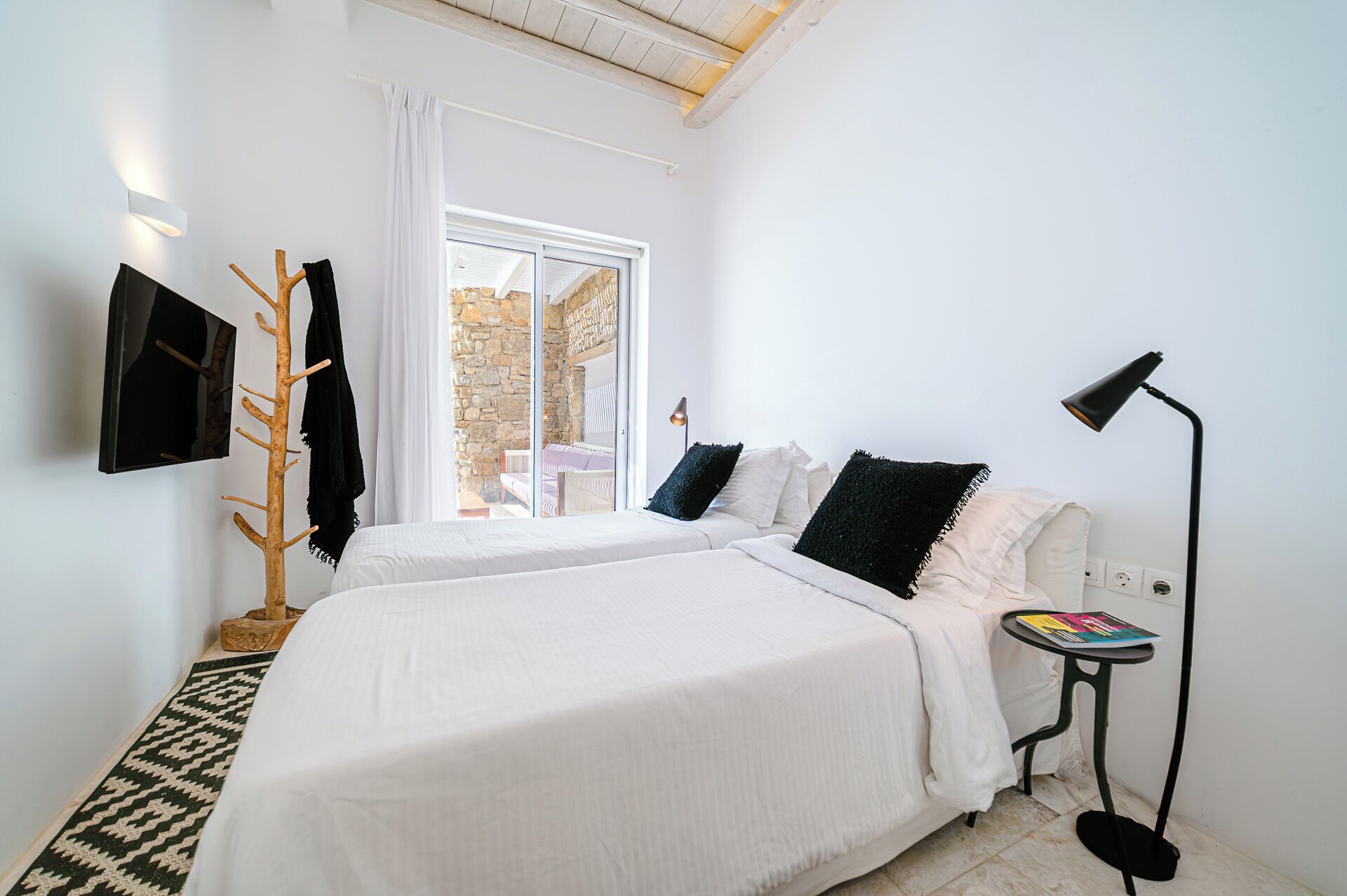 Ein Schlafzimmer mit zwei Betten, einem an der Wand montierten Fernseher, einem Holz-Kleiderständer und einem Fenster mit Blick auf ein Steinhaus.