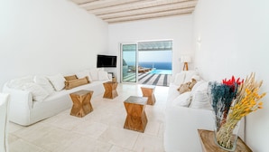 A spacious living room with a white sofa set, wooden coffee tables, and a large window offering a view of the sea.