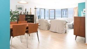Ett modernt vardagsrum med ett matbord och stolar i trä, en soffa och ett stort fönster med utsikt över havet.