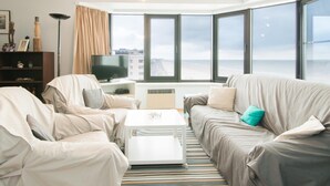 Ett modernt vardagsrum med en soffa, fåtöljer, ett soffbord och en tv. Stora fönster ger utsikt över havet.