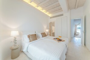 A spacious bedroom with a large bed, a bedside table with a lamp, and a view of the sea through an open door.