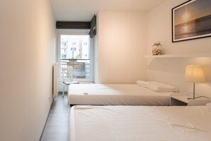 A hotel room with two beds, a bedside table with a lamp, a small shelf with decorative items, and a view of the outside balcony. - Apartment in Ostend near the Beach & Casino (OSTEND)