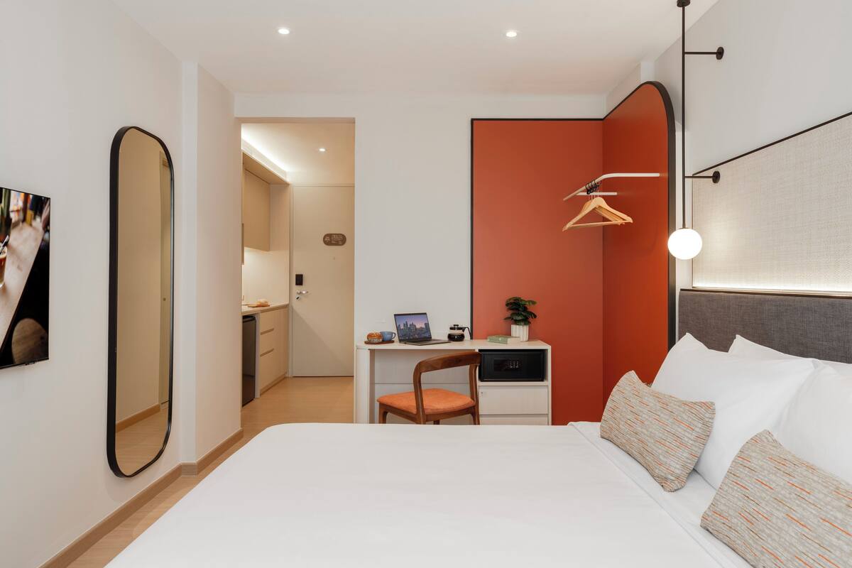 Moderni hotellihuone, jossa on suuri sänky, työpöytä kannettavalla tietokoneella, tuoli ja peili.
