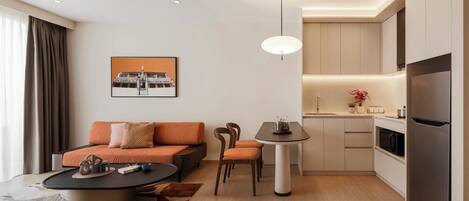 Una sala moderna con sofá, sillas, una mesa de centro y una mesa de comedor. Hay una zona de cocina con armarios, un refrigerador y un lavaplatos. Una lámpara colgante ilumina la mesa de comedor.