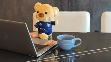 A laptop on a table with a blue mug labeled "Citadines" and a plush toy bear wearing a blue shirt with "ASCOTT" on it.