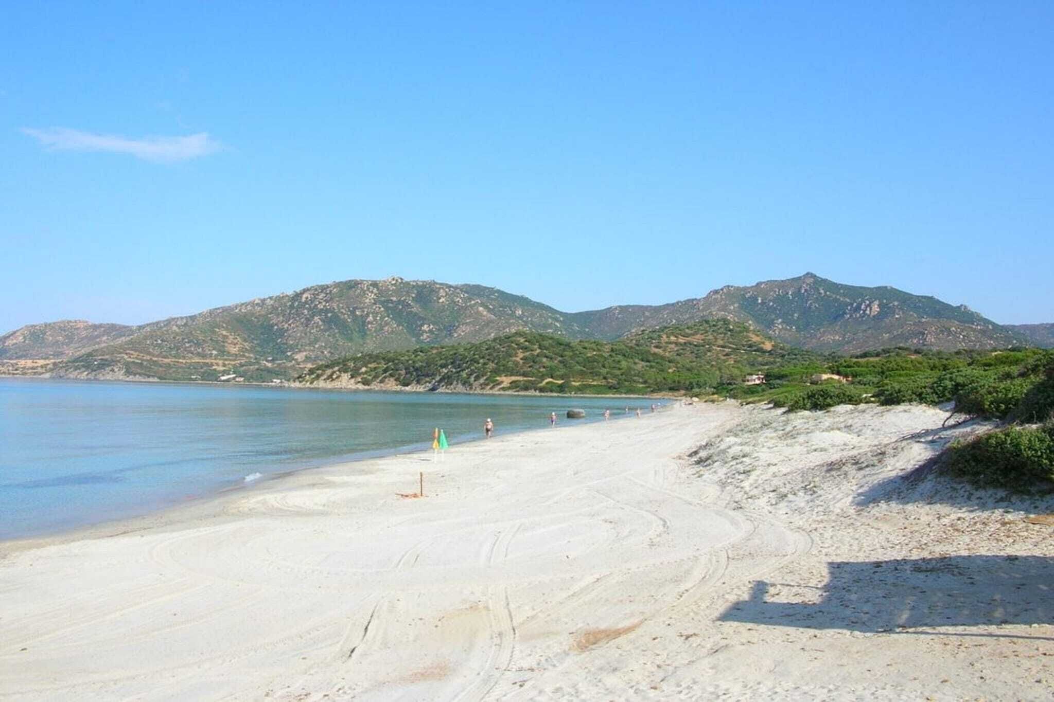 Une plage avec une eau d’un bleu limpide, un rivage sablonneux et des montagnes en arrière-plan.