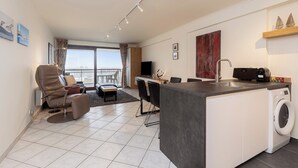 A modern living room with a sofa, a dining area, a kitchen with a washing machine, and a balcony with a view. - Apartment in Blankenberge by Marina Views (BLANKENBERGE)