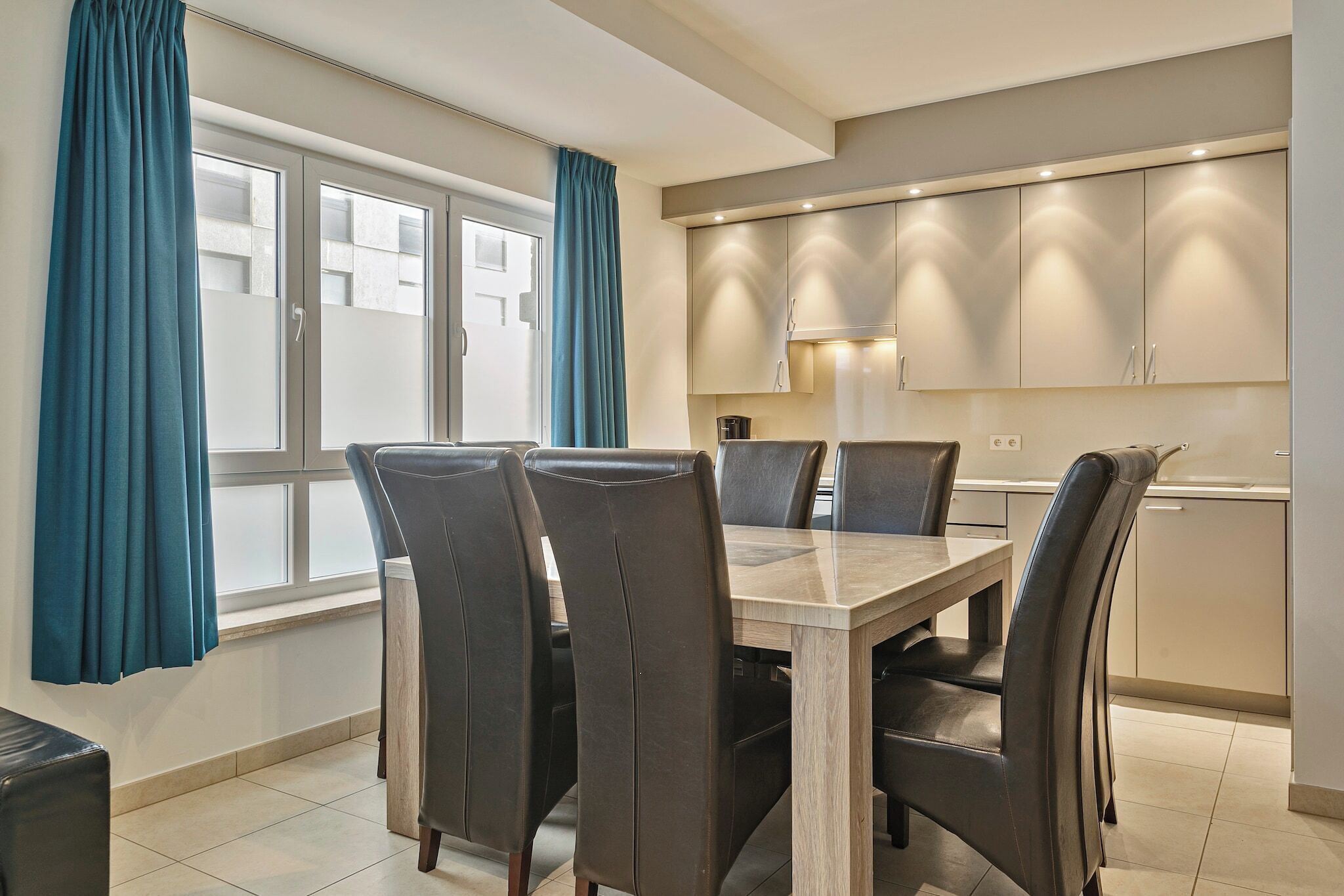 Een moderne eethoek met een rechthoekige tafel en vier stoelen, een keuken met kasten en een aanrecht, en een raam met gordijnen.