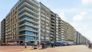 A multi-story residential building with balconies, a parking sign, and a pedestrian walkway.