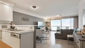 A modern kitchen with a central island, a dining area with a table and chairs, a living room with a sofa and a TV, and large windows offering a view. - Apartment in Blankenberge by the Beach (BLANKENBERGE)