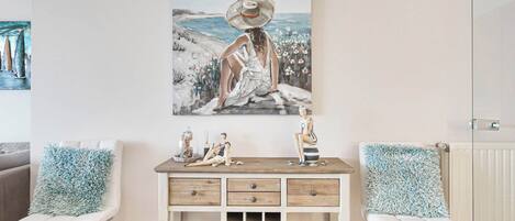 Une pièce comprenant une console en bois, deux chaises blanches avec des coussins bleus et un tableau représentant une femme au bord de la mer.