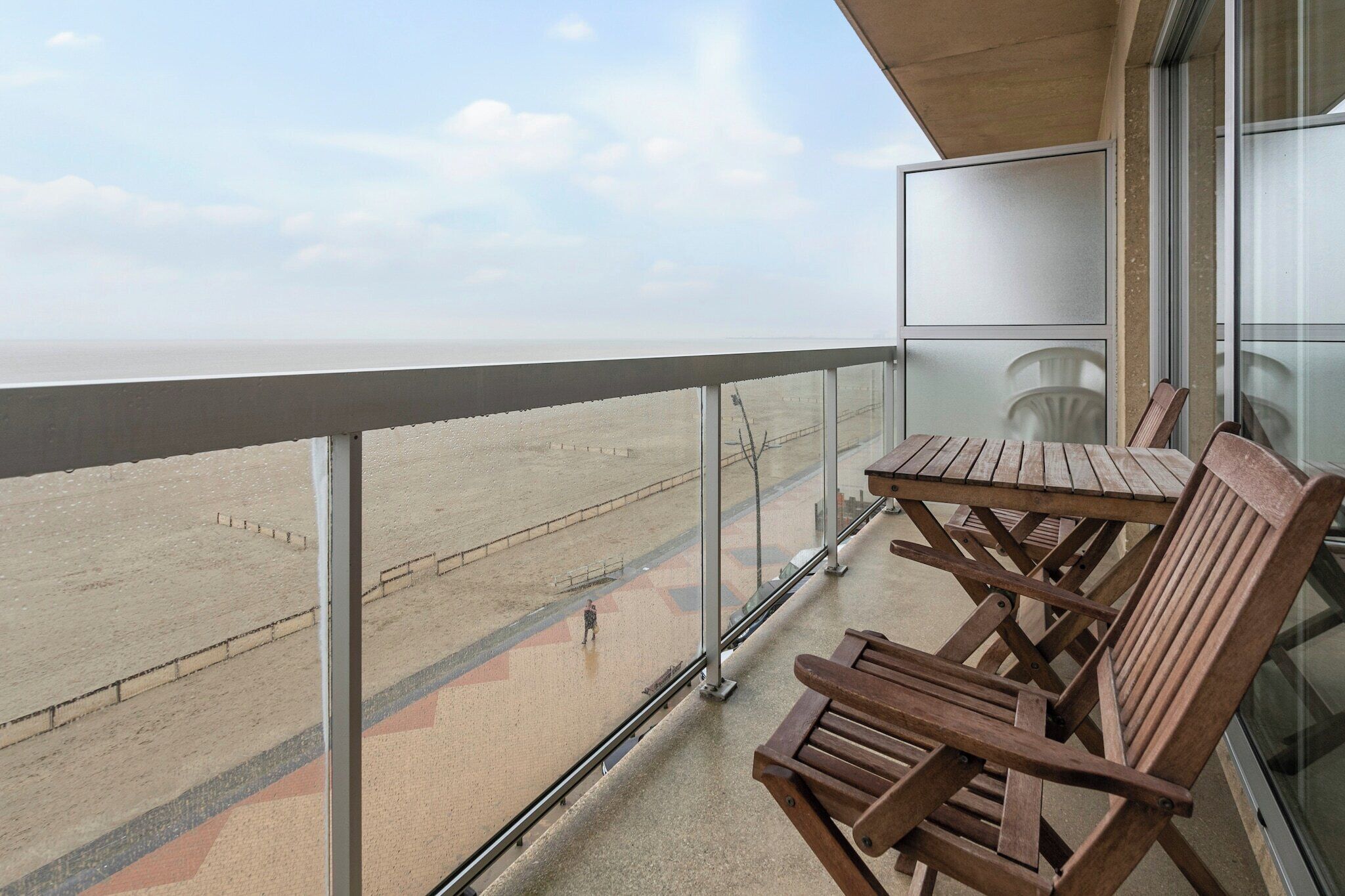 A balcony with a view of the beach, wooden chairs, and a glass railing.