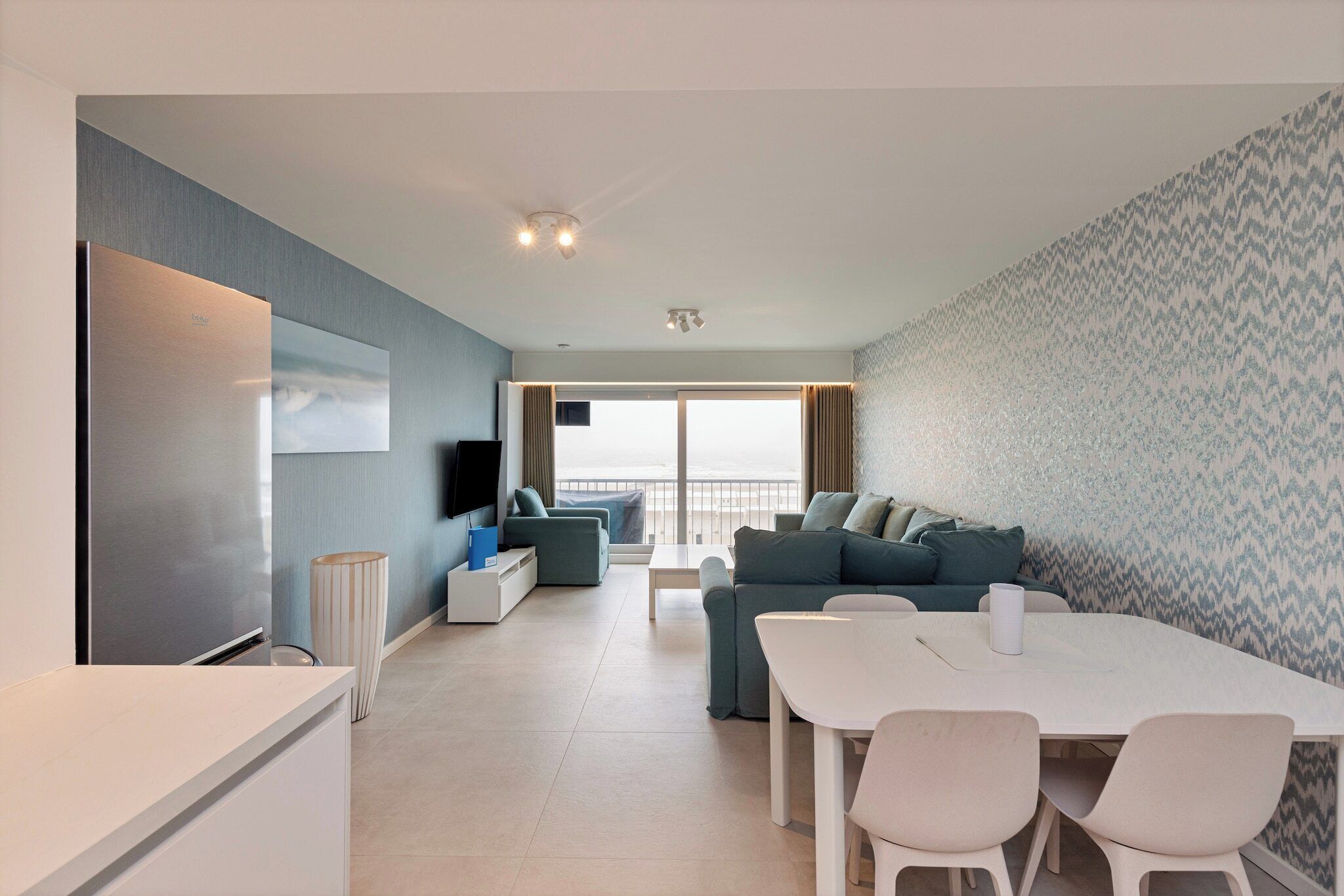 A modern living room with a flat-screen TV, a dining table with chairs, a sofa, and a large window with a view.