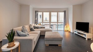 A modern living room with a grey sofa, a matching ottoman, a flat-screen TV on a white stand, and a large window with curtains. - Apartment in Blankenberge near the Beach (BLANKENBERGE)