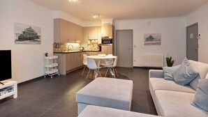 A modern living room with a grey sofa, a dining area with a white table and chairs, and a kitchen with wooden cabinets and a microwave. - Apartment in Blankenberge near the Beach (BLANKENBERGE)
