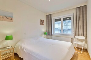 A bedroom with a bed, two bedside tables with lamps, a window with curtains, and a view of a building outside. - Flat in Blankenberge with Marina Views (BLANKENBERGE)