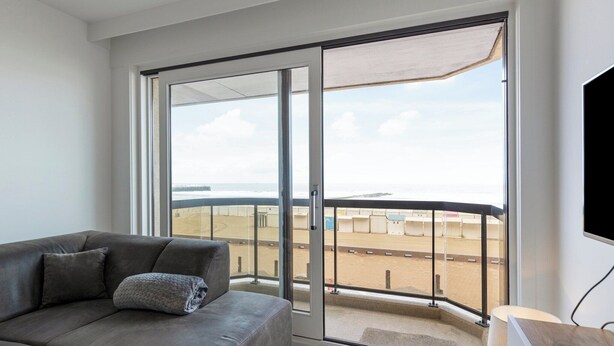 Een moderne woonkamer met een grijze bank, een grote schuifdeur naar een balkon en een flatscreen televisie aan de muur.