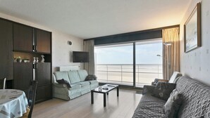 A living room with a sofa, a table, a TV, and a view of the sea. - Flat in Blankenberge near Belgium Pier (BLANKENBERGE)