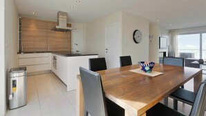 Une cuisine moderne avec des armoires blanches, un plan de travail en bois et une cuisinière intégrée. On y trouve une table à manger avec des chaises et une horloge au mur.