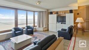 A modern living room with a large window, a sofa, a coffee table, and a TV. - Apartment by the Sea near Marina (BLANKENBERGE)
