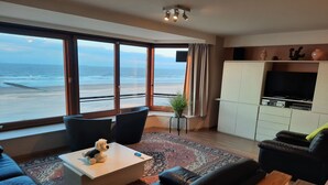 A living room with a large window overlooking the beach, a flat-screen TV, and a sofa. - Apartment by the Sea near Marina (BLANKENBERGE)