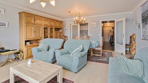 Een woonkamer met twee lichtblauwe fauteuils, een houten kast, een kroonluchter, en een eethoek met een tafel en stoelen.