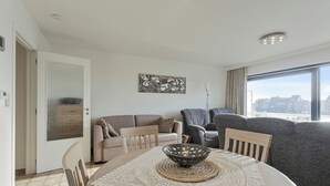 A modern living room with a dining table, two sofas, a large window with a city view, and a ceiling light fixture. - Apartment in Blankenberge near Belgium Pier (BLANKENBERGE)