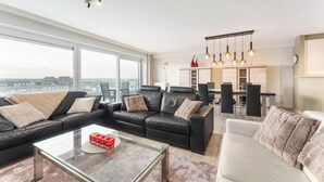 A modern living room with a glass coffee table, black leather sofas, and a white sofa. There is a dining area with a table and chairs, and a kitchen in the background. - Apartment in Blankenberge near Belgium Pier (BLANKENBERGE)