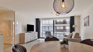 Una sala de estar moderna con una mesa redonda de madera, un televisor de pantalla plana y un ventanal con vistas a un edificio.