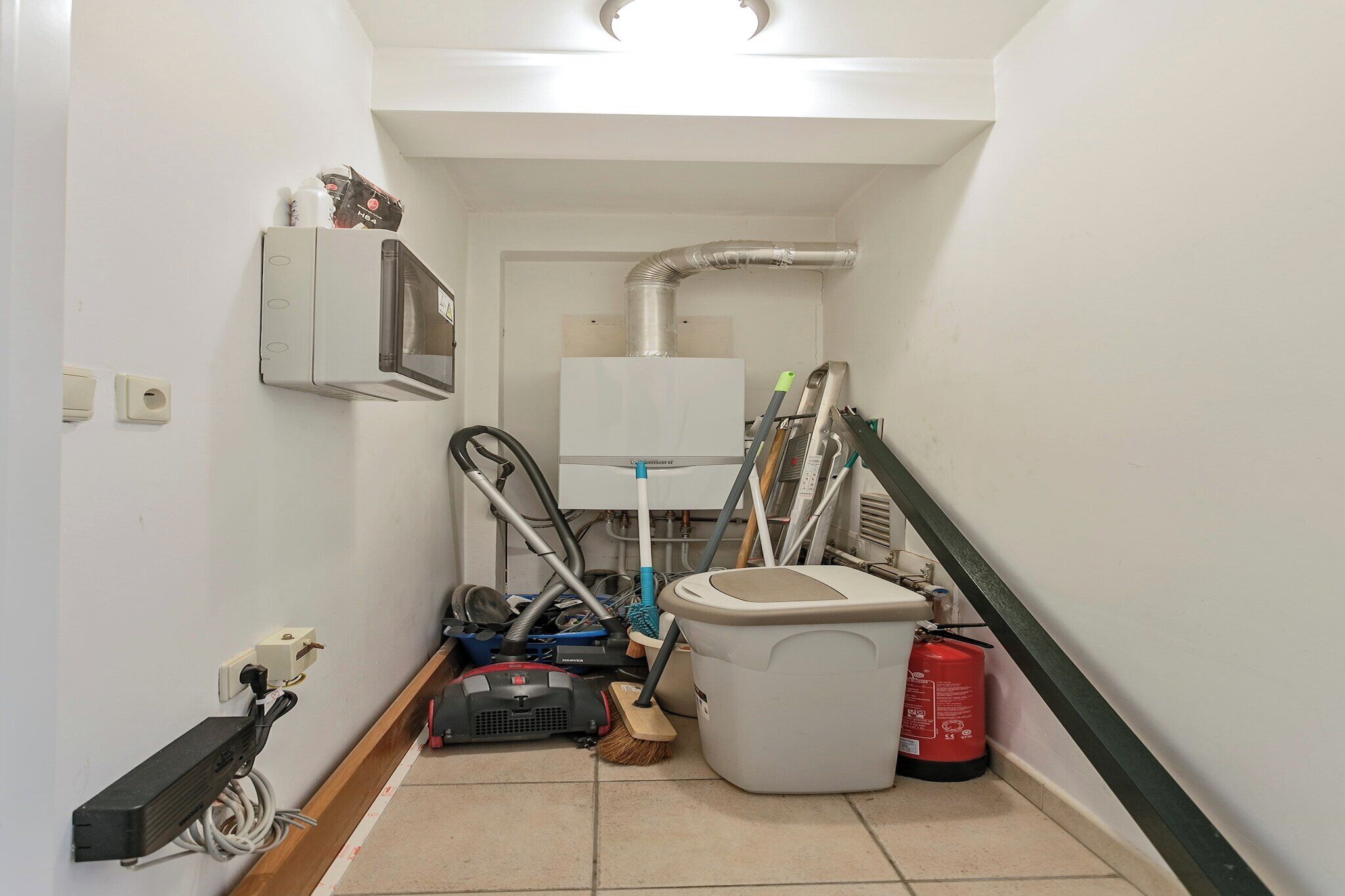A narrow hallway with a white wall, a white toilet, a red fire extinguisher, a vacuum cleaner, and a broom.
